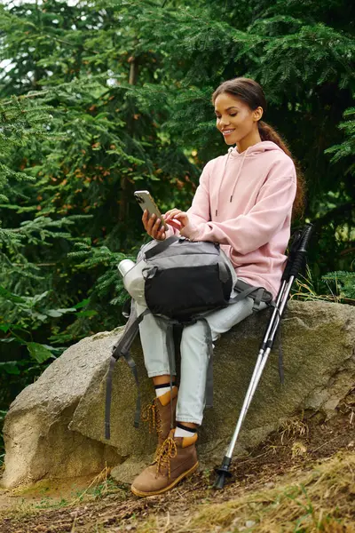 A young African American woman sits on a rock, enjoying her forest hike and checking her phone amidst autumn colors. — Stock Photo