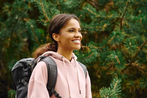 A young African American woman smiles brightly while hiking through a colorful autumn forest trail. — Stock Photo