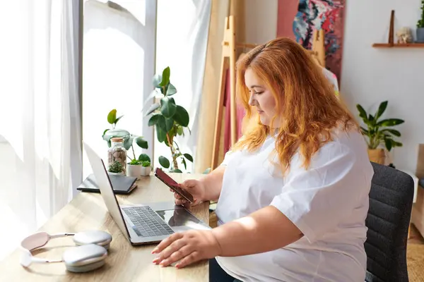 A plus size woman sits at her desk, absorbed in her phone, surrounded by a warm, inviting space. — Stock Photo