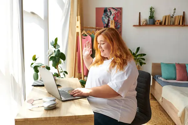 A joyful plus size woman smiles while waving during a video call at her cozy home office. — Stock Photo