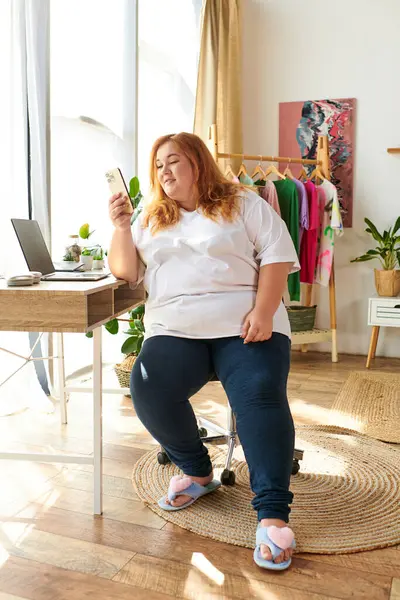 Веселая женщина plus size наслаждается своим временем дома, используя свой телефон, сидя за уютным столом. — стоковое фото