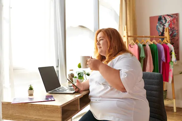 A vibrant plus size woman sips coffee while working on her laptop, surrounded by colorful clothes. — Stock Photo
