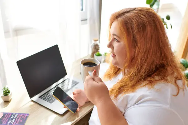 Una bella donna plus size sorseggia caffè e controlla il suo telefono in uno spazio di lavoro luminoso e allegro. — Foto stock
