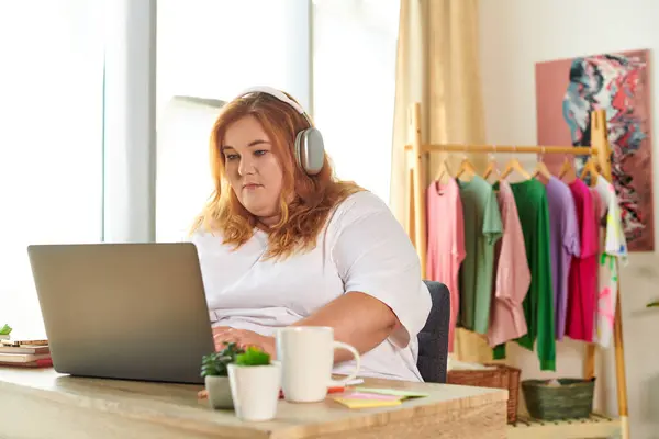 A plus size woman engages with her laptop, headphones on, in a colorful, cozy workspace. — Stock Photo