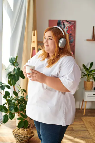 Eine fröhliche Plus-Size-Frau entspannt bei einem Drink, Kopfhörer auf, umgeben von Zimmerpflanzen. — Stockfoto