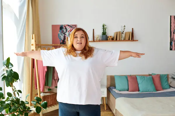 A plus size woman smiles while standing with arms open wide, celebrating her body in a warm space. — Stock Photo
