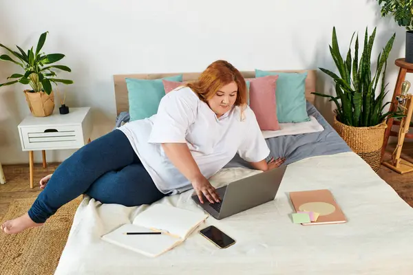 Уверенная в себе женщина plus size занимается со своим ноутбуком, уютно сидя в яркой спальне. — стоковое фото