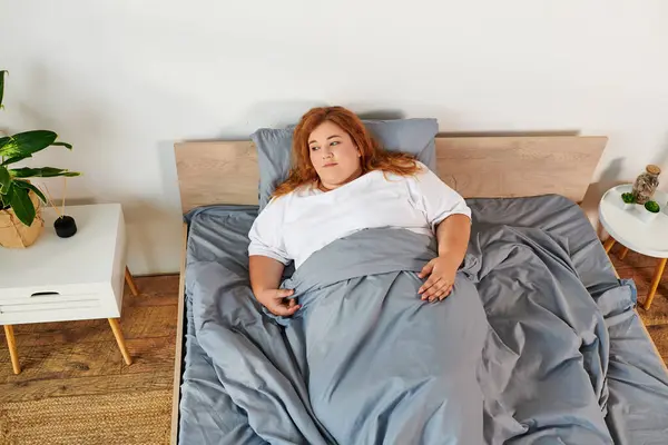 A lovely plus size woman lounges peacefully in bed, wrapped in soft gray covers. — Stock Photo