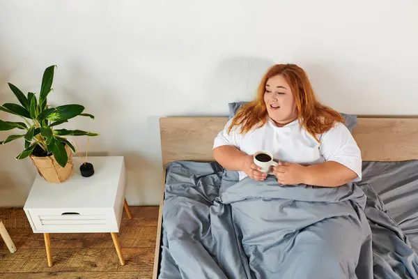 Una mujer alegre de tamaño grande se relaja en la cama con una taza de café caliente, abrazando la luz de la mañana. - foto de stock