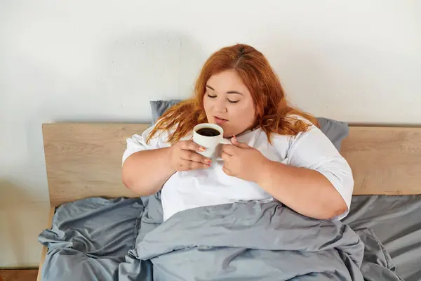 Una encantadora mujer de tamaño grande sorbe café envuelto en mantas acogedoras, disfrutando de su mañana tranquila. - foto de stock