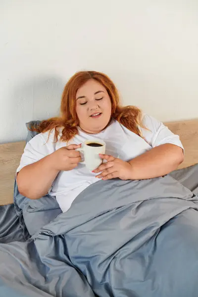 Eine schöne Plus-Size-Frau genießt eine warme Tasse Kaffee, während sie sich in ihr gemütliches Bett schmiegt. — Stockfoto