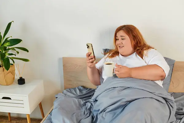 A beautiful plus size woman lounges in bed, sipping coffee and checking her phone. — Stock Photo