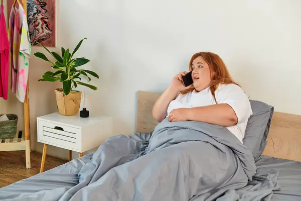 Eine schöne Plus-Size-Frau genießt einen Telefonanruf, eingewickelt in weiche Bettwäsche, umgeben von Wohnkultur. — Stockfoto