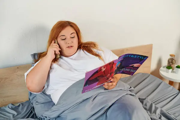 Eine schöne Frau führt Gespräche, während sie in einem Magazin blättert und es sich in ihrem Bett gemütlich macht. — Stockfoto