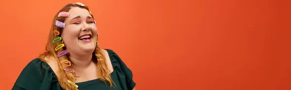 A joyful plus size woman laughs, flaunting colorful hair clips by an orange wall. — Stock Photo