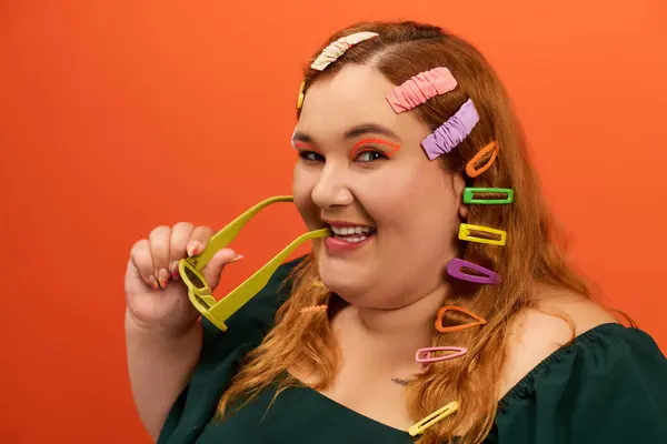 A cheerful woman with a bright smile playfully holds green glasses amid colorful clips. — Stock Photo