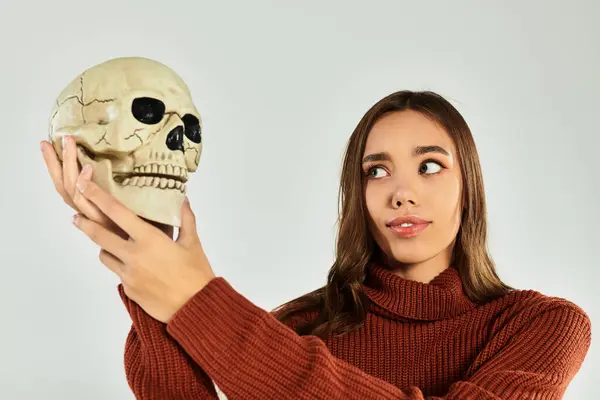A young, beautiful woman in a sweater holds a skull, embodying Halloween vibes with curiosity. — Stock Photo