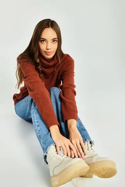 A beautiful young woman sits on the floor, dressed in a stylish sweater and jeans, radiating warmth. — Stock Photo
