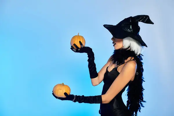 Vestida com um traje de bruxa impressionante, uma jovem mulher equilibra elegantemente duas abóboras ao posar. — Fotografia de Stock