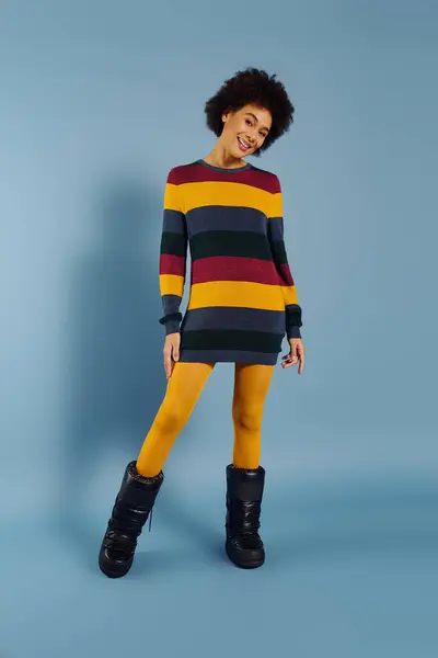 A joyful young woman in a colorful striped sweater and yellow leggings shines in a bright space. — Stock Photo