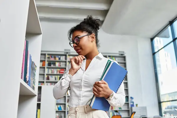 A thoughtful teacher examines books in a modern library setting, surrounded by shelves and light. — Stock Photo