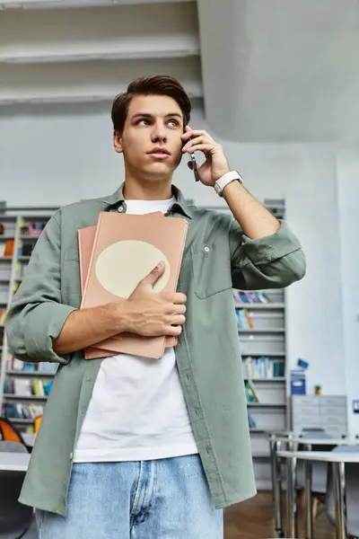 Un estudiante dedicado está parado en una biblioteca, sosteniendo libros y hablando por teléfono intensamente. - foto de stock