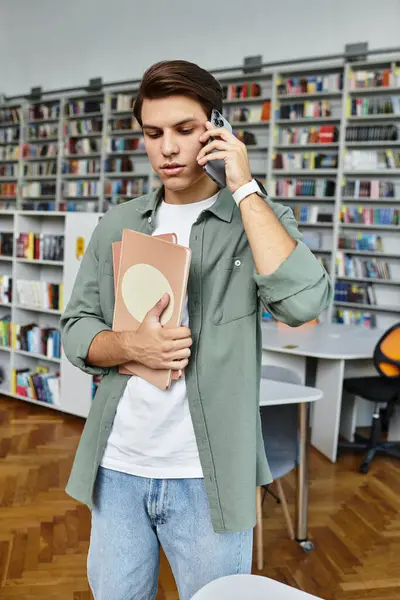 A focused young man holds books and speaks on the phone, immersed in study at his campus library. — Stock Photo