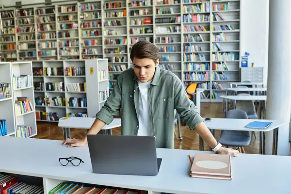 Um estudante focado se envolve com seu laptop em meio a prateleiras de livros em uma biblioteca. — Fotografia de Stock