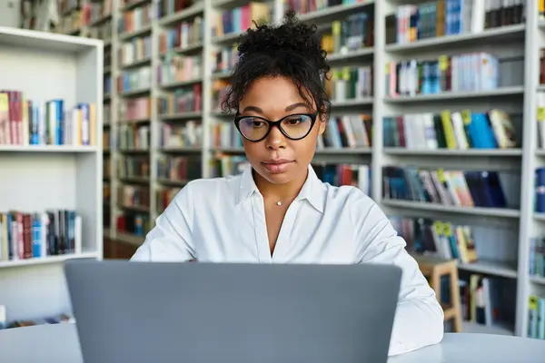 Un'insegnante dedicata recensisce i materiali sul suo computer portatile mentre è circondata da scaffali pieni di libri. — Foto stock