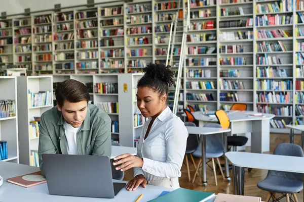 A teacher engages a young student while they explore a laptop amidst shelves of books. — Stock Photo