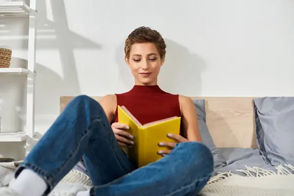 С скрещенными ногами на кровати, молодая женщина читает ярко-желтую книгу в мирный день дома. — стоковое фото