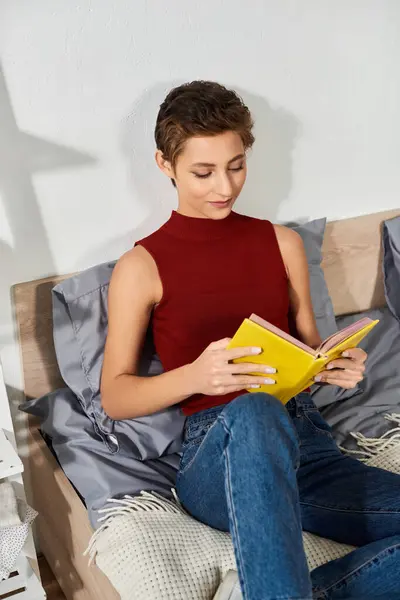 Una giovane donna con i capelli corti e trucco naturale si rilassa a casa, assorta in un libro durante la sua routine. — Foto stock