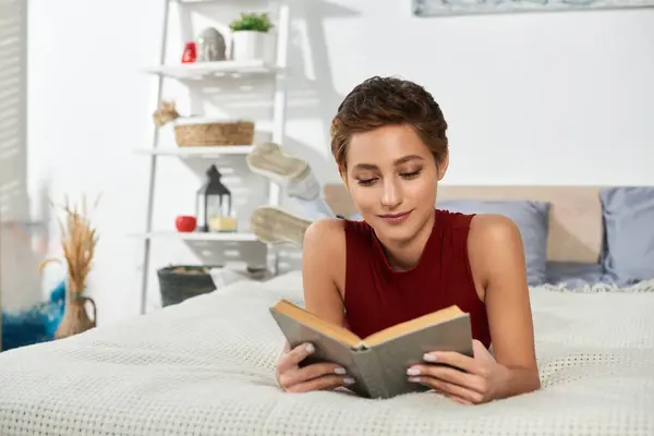 Молодая женщина расслабляется дома, погружается в хорошую книгу, одеваясь комфортно в красную майку. — стоковое фото