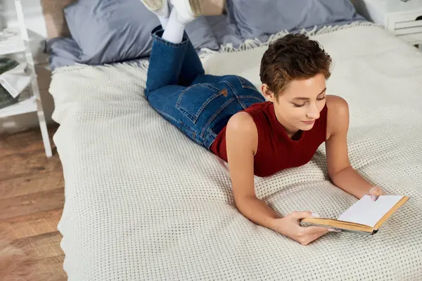 Entspannt zu Hause liest eine junge Frau im roten Tank-Top ein Buch, während sie es sich auf ihrem Bett bequem macht. — Stockfoto