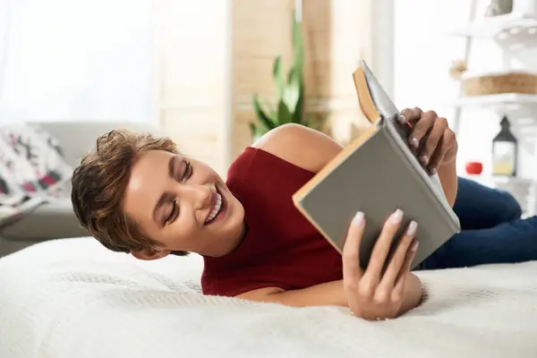 A joyful young woman enjoys a quiet moment at home, immersed in a good book while lying on her bed. — Stock Photo