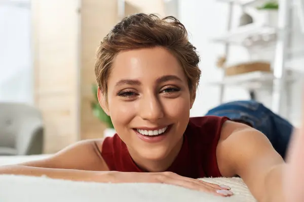 A young woman with short hair smiles warmly while lounging at home, embracing a relaxed and happy moment. — Stock Photo
