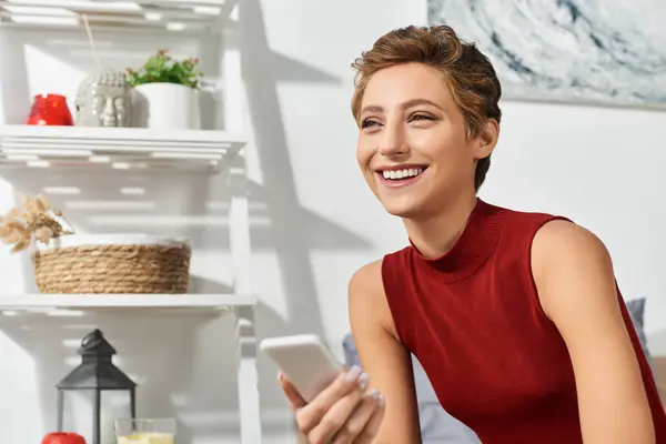 Une jeune femme joyeuse dans un débardeur rouge se détend à la maison tout en s'engageant avec son téléphone, rayonnant de joie. — Photo de stock