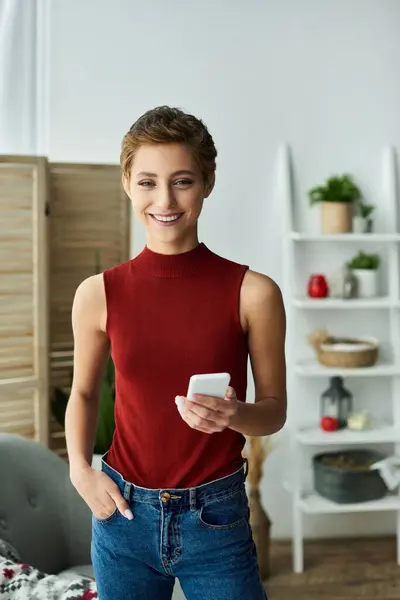 A young woman in a red tank top smiles warmly at home as she engages with her smartphone. — Stock Photo