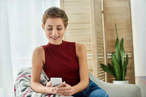Bright sunlight fills the cozy space as a young woman relaxes at home, smiling while using her phone. — Stock Photo