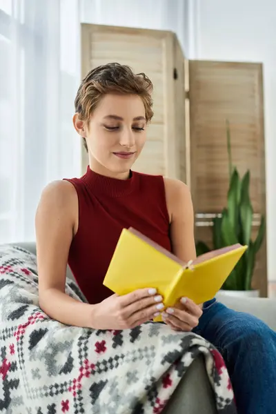 A young woman in a red tank top relaxes at home, absorbed in her yellow book while embracing her casual routine. — Stock Photo