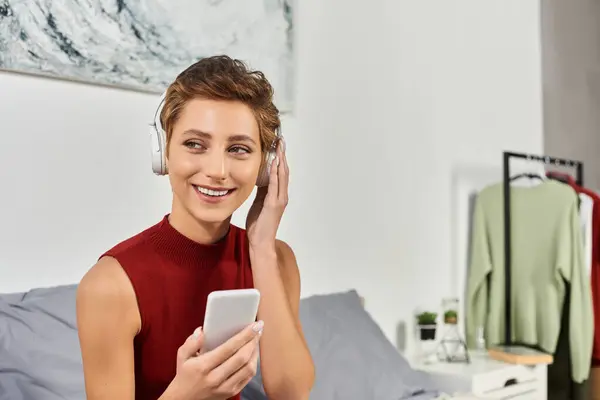 A young woman with short hair relaxes at home, listening to music and smiling in her red tank top. — Stock Photo