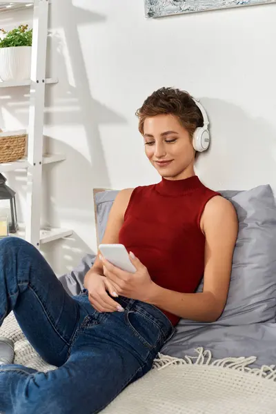 A young woman relaxes at home, wearing a red tank top and headphones, absorbed in her smartphone. — Stock Photo