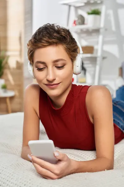 A young woman in a red tank top lounges at home, enjoying music and scrolling through her phone content. — Stock Photo
