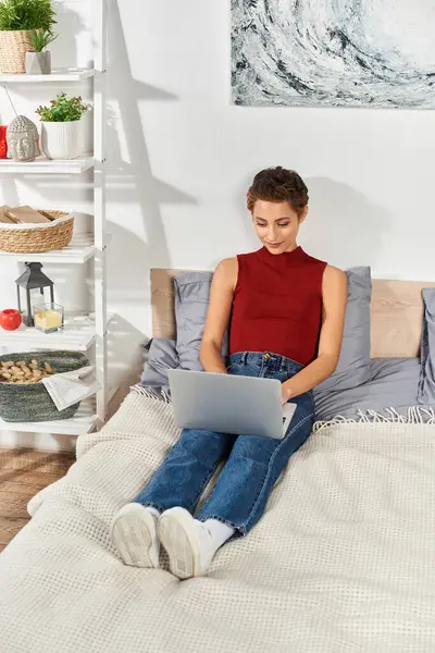 Relaxing at home, a young woman in a red tank top focuses on her laptop, enjoying her daily routine. — Stock Photo