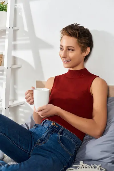 A young woman in a red tank top sips coffee while lounging at home, embracing the peaceful daily routine. — Stock Photo