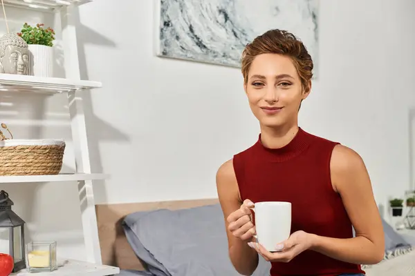 Relaxante em casa, uma jovem mulher em um top tanque vermelho saboreia seu café da manhã, abraçando um momento de paz. — Fotografia de Stock