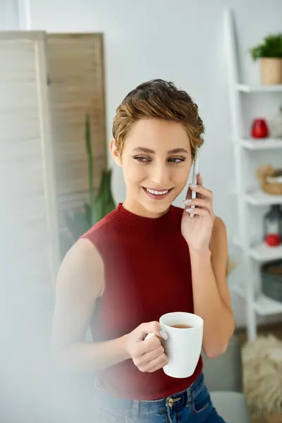 Una joven en un tanque rojo sonríe mientras habla por teléfono, sosteniendo una taza de café en casa. - foto de stock