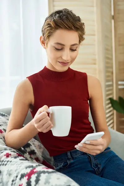 Una joven con el pelo corto se relaja en casa, bebiendo café y centrada en su teléfono en un ambiente acogedor. - foto de stock