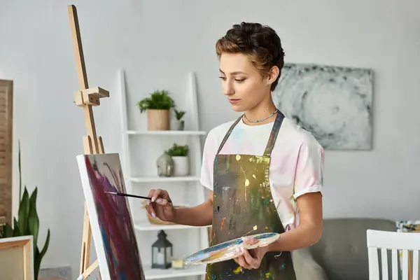 A young woman with short hair focuses intently as she paints on a canvas in her artistic home space. — Stock Photo