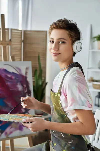 Una joven con maquillaje natural inmersa en la pintura expresa felizmente su creatividad en una acogedora habitación. - foto de stock